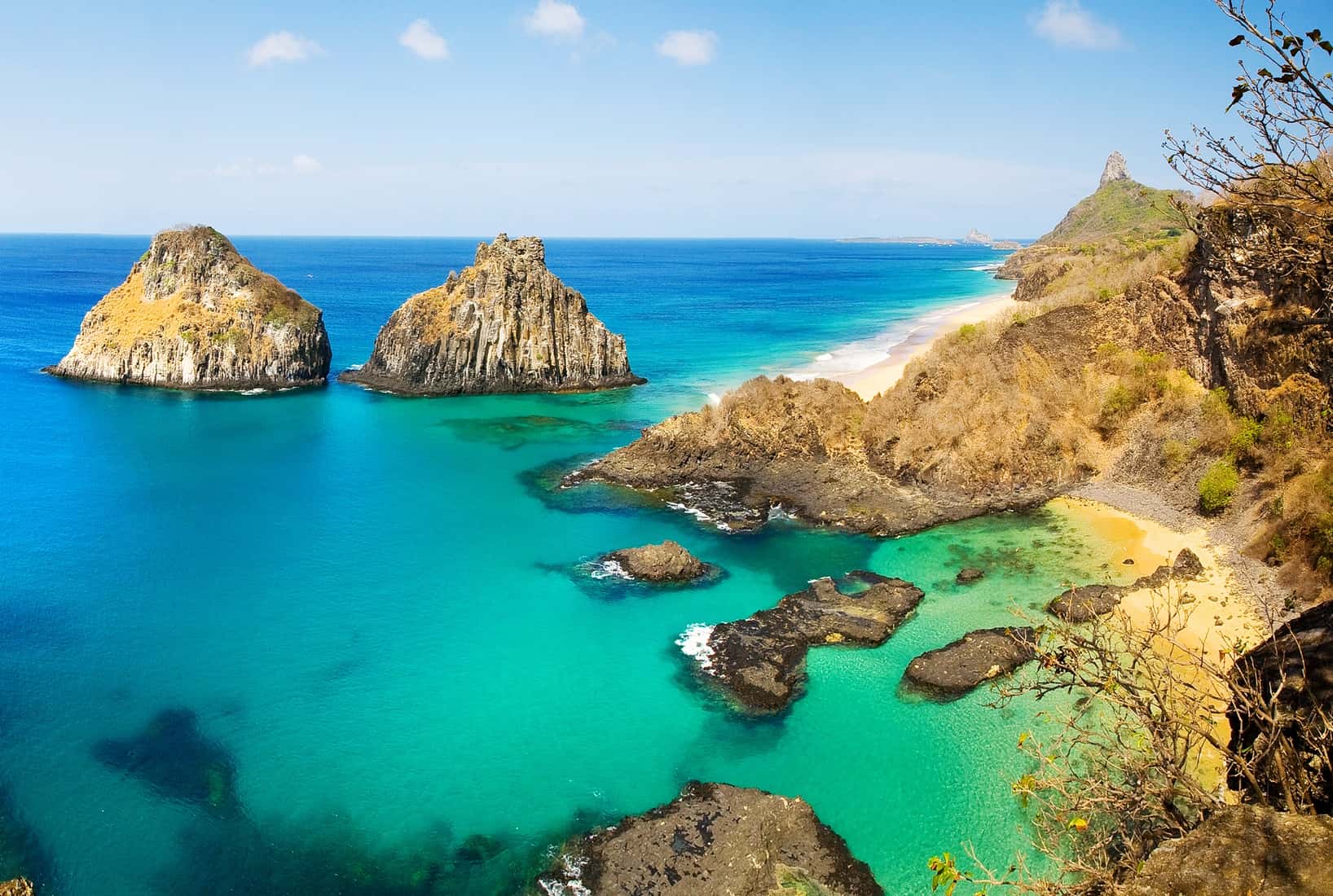 Baía dos Porcos em Fernando de Noronha com águas azul-turquesa, falésias vulcânicas e os Dois Irmãos ao fundo.