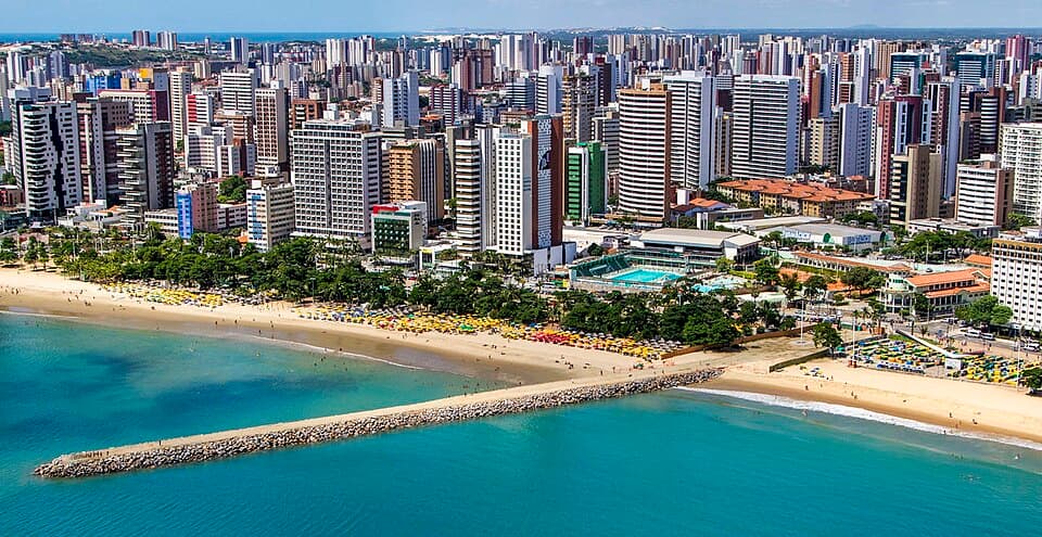 Orla de Fortaleza com praia de areia dourada, mar azul, coqueiros e skyline da cidade ao fundo.